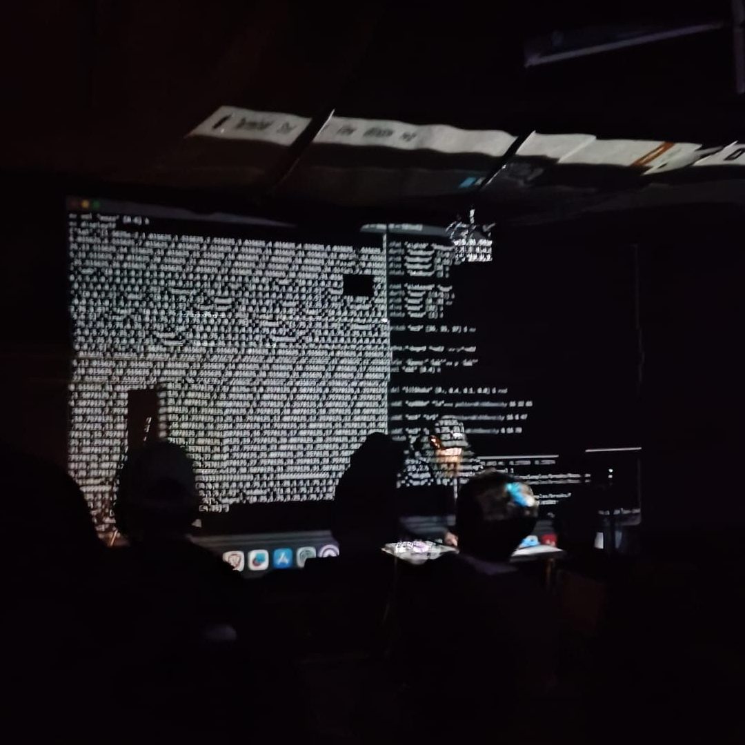 The image shows a person playing a live coding performance in a dimly lit room with code being projected on the wall behind him and on the person himself
